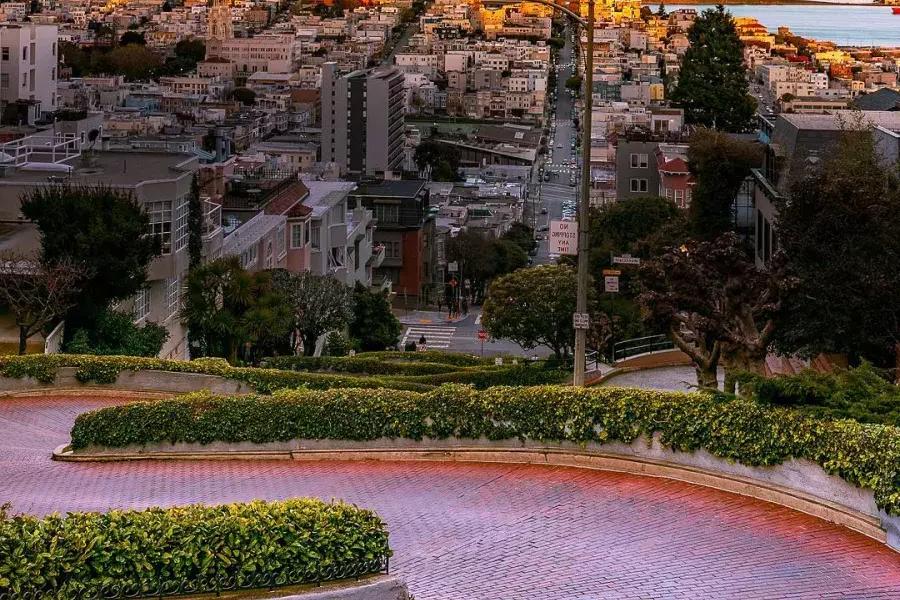 Lombard Street) curves with Coit Tower in the distance during sunset.
