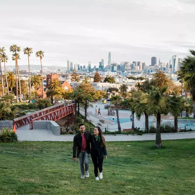 ドロレス公園とサンフランシスコのスカイラインを背にカメラに向かって歩くカップル。
